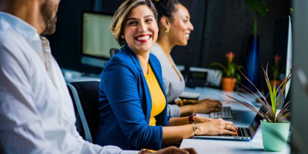Three diverse professionals working and smiling at office desks, fostering teamwork and collaboration.