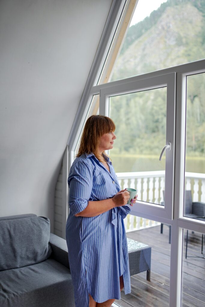 Dreamy middle aged female in casual wear with cup of hot drink contemplating mountain from countryside house while looking away