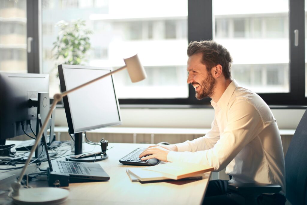 7 Proven Ways to Make Money Online in 2025 (Beginners Friendly) 2 A man smiling while working at an office desk with a computer and natural daylight streaming in through large windows.